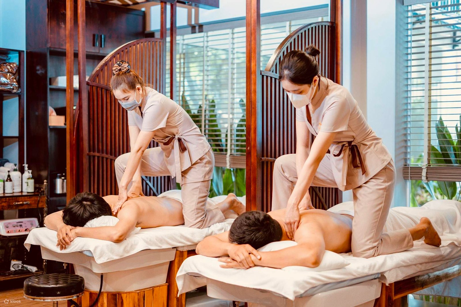 Hai người đang được massage ở spa, với nhân viên đeo khẩu trang thực hiện liệu pháp trên giường massage.