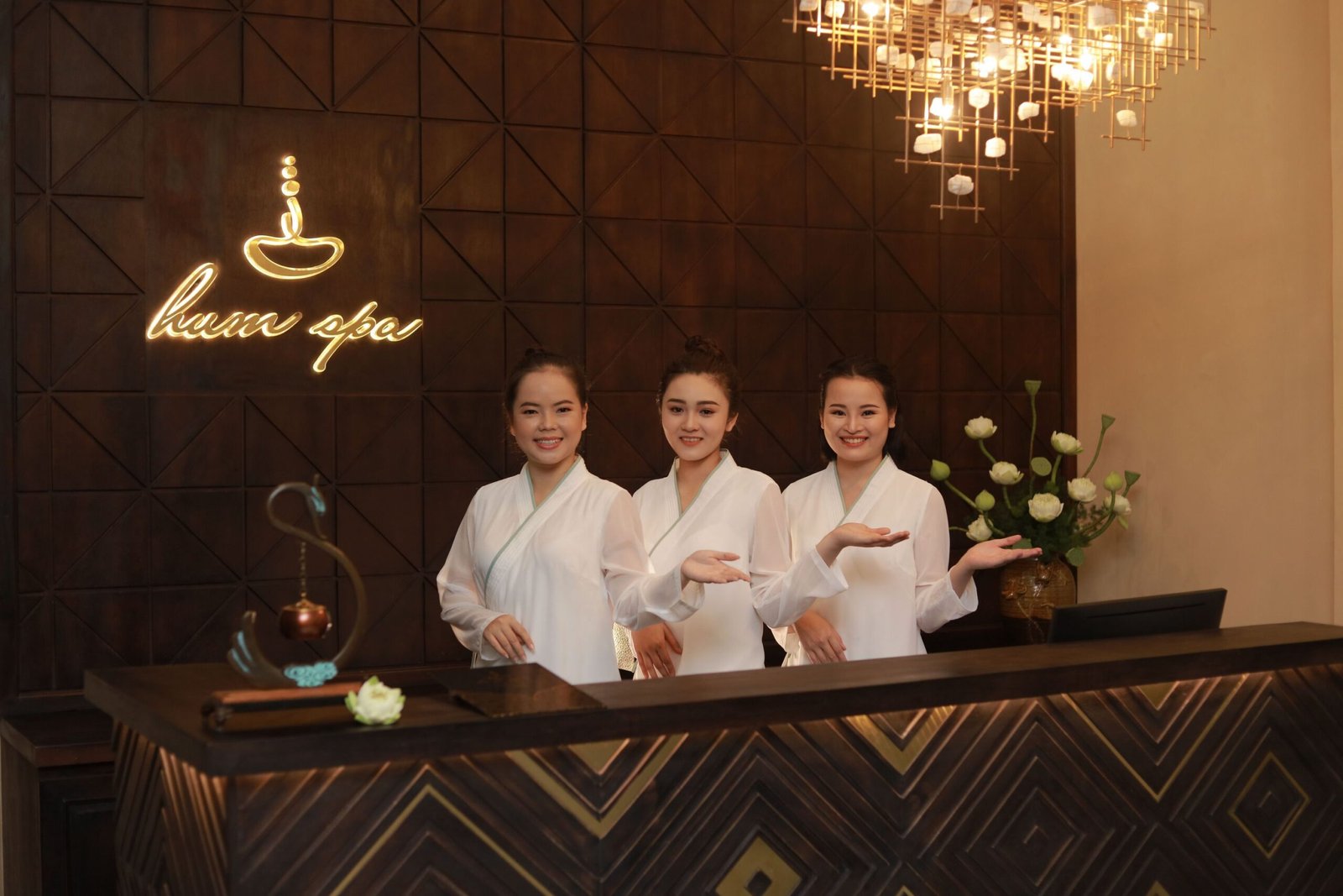 Ba nhân viên mặc áo trắng đứng sau quầy lễ tân spa với nụ cười rạng rỡ, trong ánh đèn ấm áp.