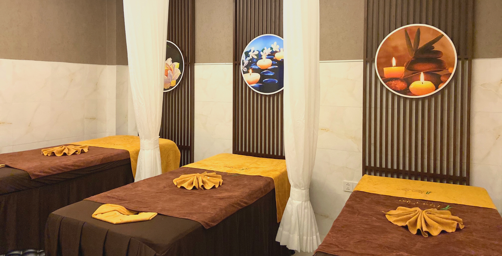 Ba giường massage với khăn trải màu nâu và vàng, trang trí tranh tường với nến và đá, tạo không gian thư giãn.