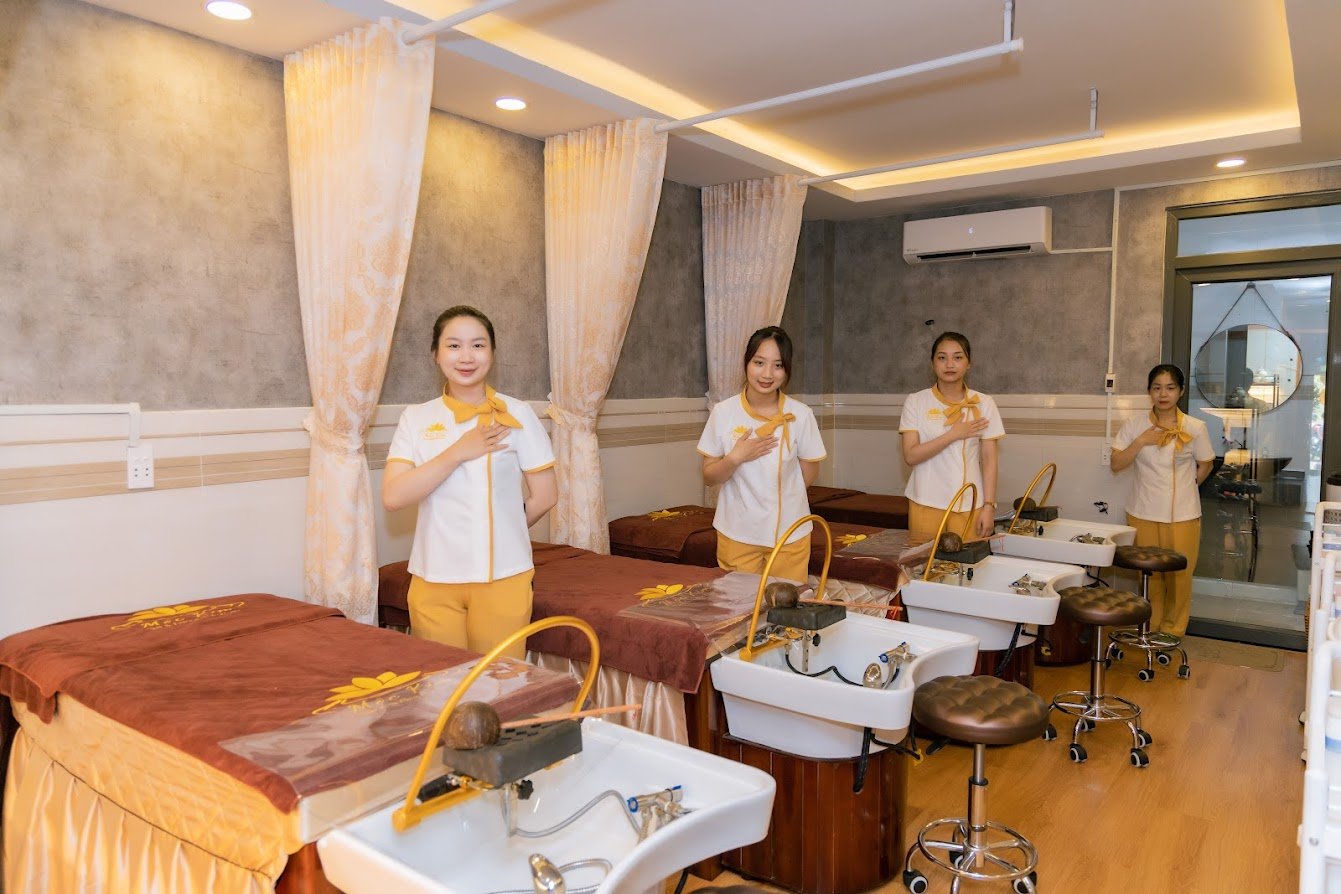 Nhân viên trong đồng phục đứng trong phòng spa với giường massage và bồn rửa, ánh sáng ấm áp.
