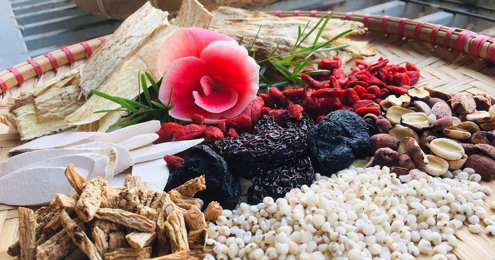 Các loại thảo dược và hạt khô được sắp xếp trên mẹt tre, có hoa hồng trang trí ở giữa.