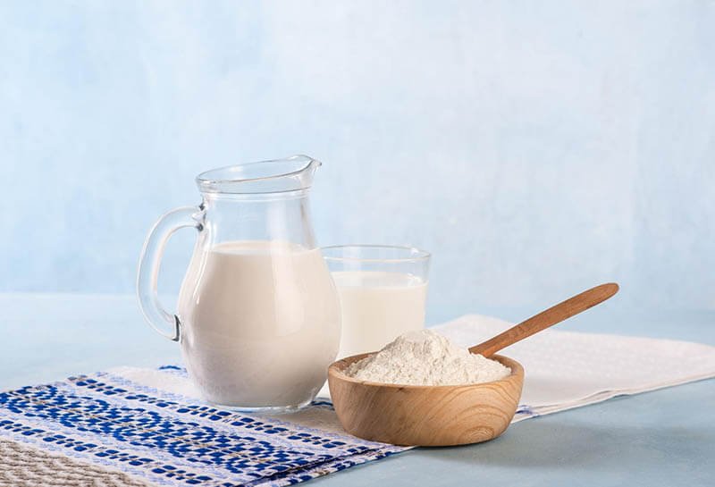 Bình sữa, ly sữa và bát bột trên khăn trải bàn xanh trắng, nền xanh nhạt.