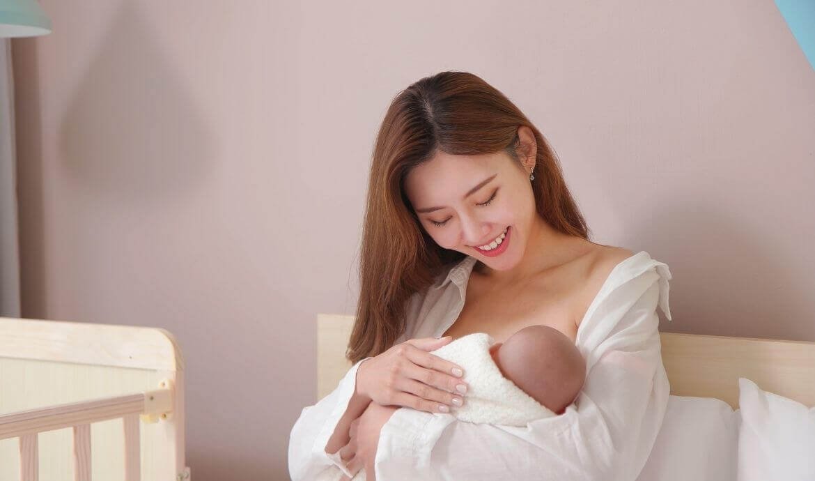 Người phụ nữ mỉm cười, bế em bé trong vòng tay, ngồi cạnh nôi trẻ em, trong không gian ấm áp.