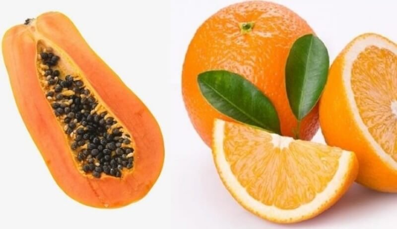 Đu đủ cắt đôi với nhiều hạt đen bên trong, bên cạnh là cam nguyên trái và cam cắt lát, kèm lá xanh.