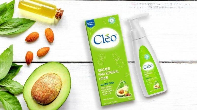 Hình ảnh chai và hộp kem tẩy lông Cléo, bên cạnh là quả bơ, lá cây, hạnh nhân và lọ dầu trên nền trắng.