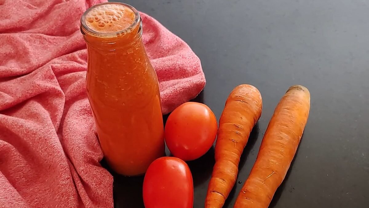 Một chai nước ép màu cam, hai củ cà rốt và hai quả cà chua trên nền vải đỏ và mặt bàn đen.