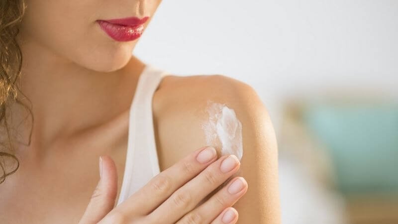 Người phụ nữ thoa kem dưỡng da lên vai, mặc áo trắng và tô son đỏ, nền mờ với màu xanh và nâu.