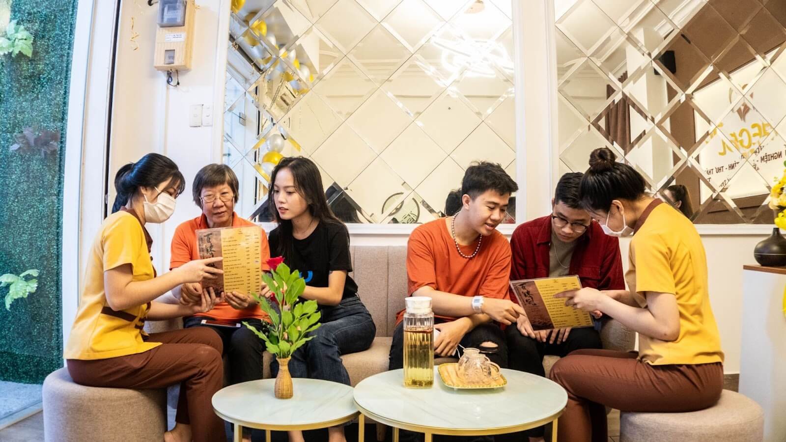 Nhóm người đang ngồi trong quán cà phê, xem thực đơn, trước bàn có bình hoa và đồ uống.