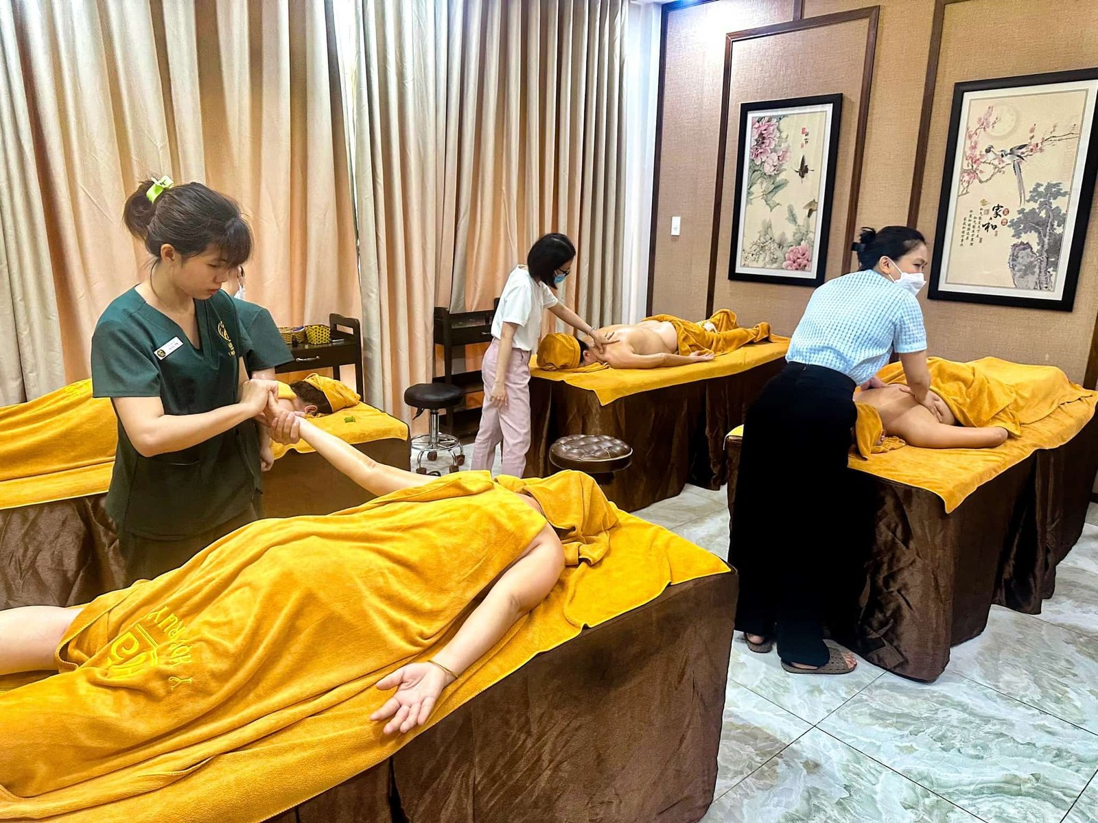 Nhân viên spa đang thực hiện massage cho khách hàng trên giường, trong phòng có trang trí tranh treo tường.