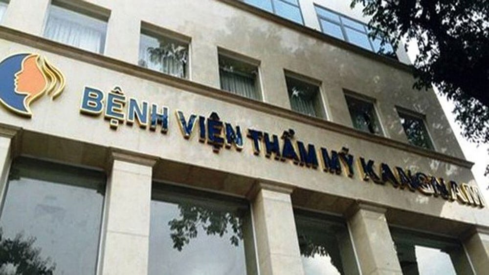 Tòa nhà với biển hiệu "Bệnh viện Thẩm mỹ Kangnam", nền trời và tán cây bên góc phải.