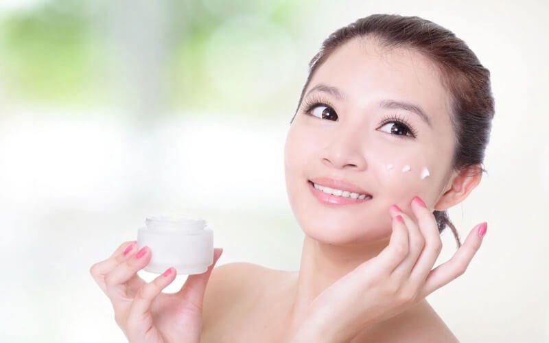 Người phụ nữ cầm hũ kem, mỉm cười và thoa kem lên má trong bối cảnh sáng tự nhiên.