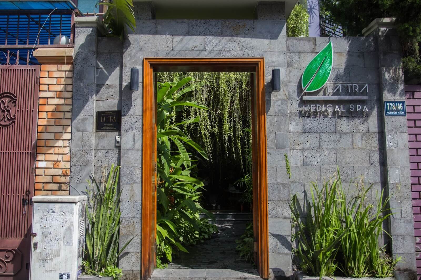 Lối vào một spa với cổng đá xám, cây xanh bao quanh, biển hiệu có hình chiếc lá màu xanh.