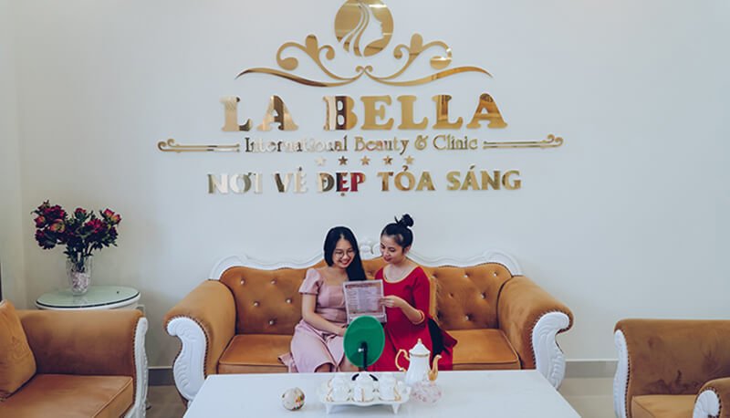 Hai người ngồi trên sofa tại thẩm mỹ viện, cùng xem tài liệu. Không gian ấm cúng và sang trọng.