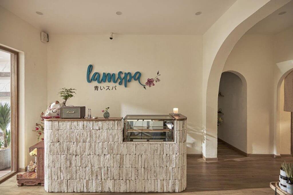Quầy lễ tân spa với tường gạch trắng, trang trí bằng cây xanh và nến, có biển hiệu "lamspa".