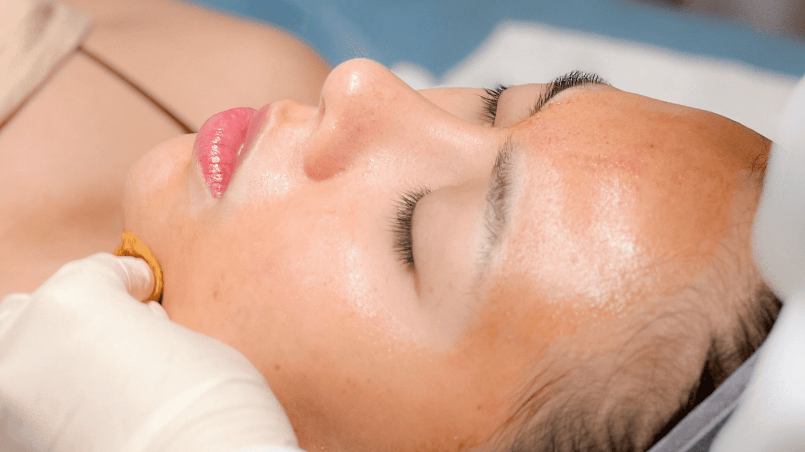 Người đang nằm nhắm mắt, nhận chăm sóc da mặt với tay đeo găng tay đang thực hiện thao tác.