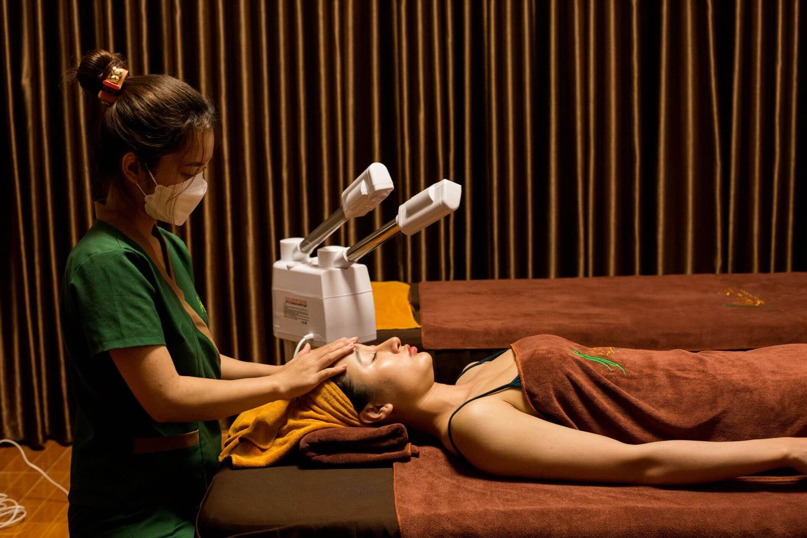 Người đang tận hưởng thư giãn trên giường massage trong không gian spa ấm áp với rèm nâu và ánh đèn dịu.