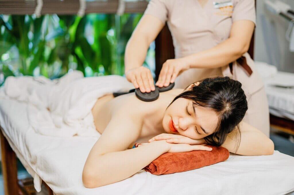 Người đang tận hưởng liệu pháp mát-xa với đá nóng trên lưng, nằm thư giãn trên giường trong không gian spa.