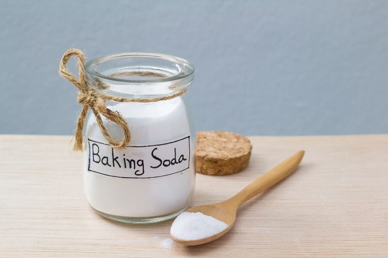 Hũ chứa bột baking soda với nắp đậy và muỗng gỗ trên mặt bàn gỗ sáng.