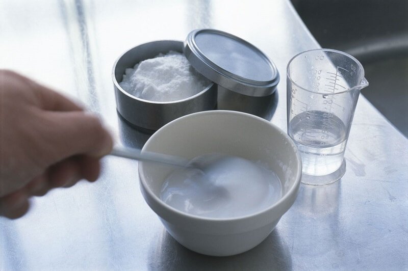 Người đang khuấy hỗn hợp trong bát, cạnh đó là lọ bột và cốc nước trên bàn kim loại.