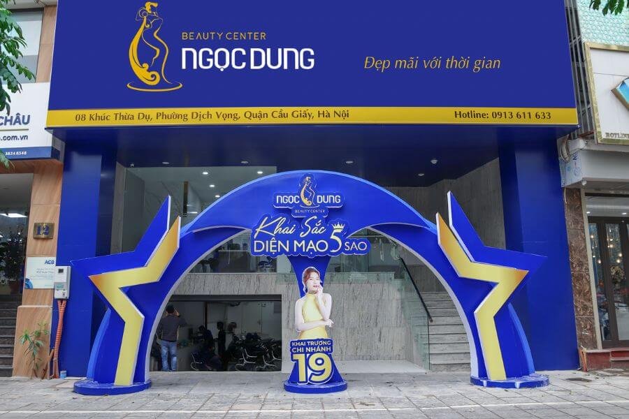 Mặt tiền trung tâm làm đẹp với biển hiệu màu xanh, có cổng chào và bức ảnh quảng bá chi nhánh mới.