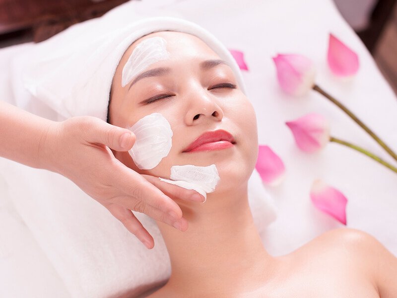 Người phụ nữ thư giãn trong spa với mặt nạ dưỡng da trên mặt, xung quanh có cánh hoa màu hồng.
