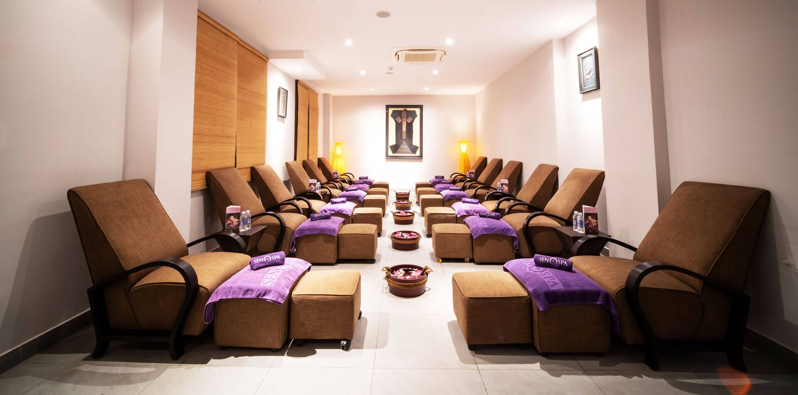 Phòng spa với ghế thoải mái xếp thành hàng, khăn màu tím và chậu nước hoa bên dưới, ánh sáng ấm áp.