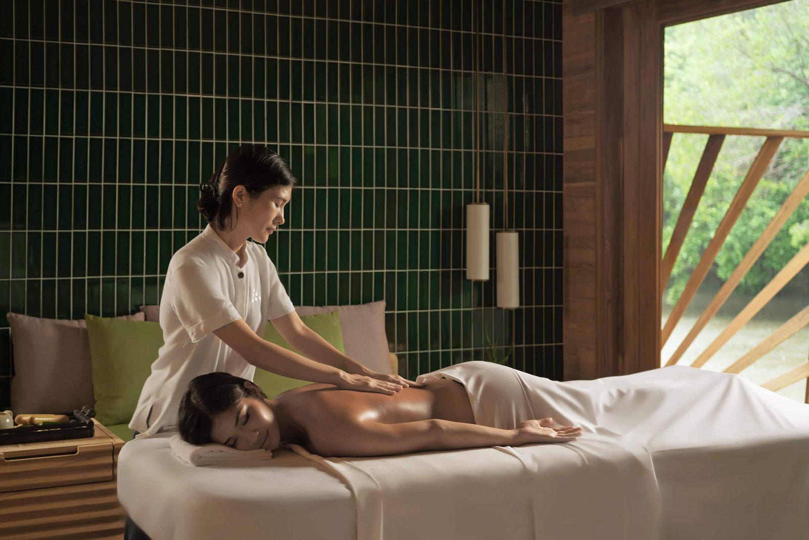 Người đang thư giãn trên giường massage, trong căn phòng có tường gạch xanh và cửa sổ lớn, ánh sáng dịu nhẹ.