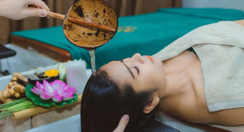 Người đang nằm thư giãn trong spa, có nước được đổ lên đầu, xung quanh có hoa sen và nến trang trí.