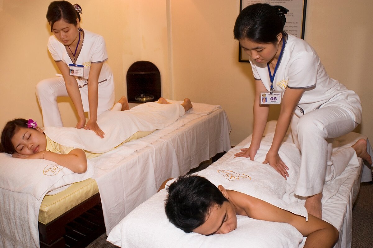 Hai người đang được massage thư giãn trên giường, với nhân viên massage đứng bên cạnh.