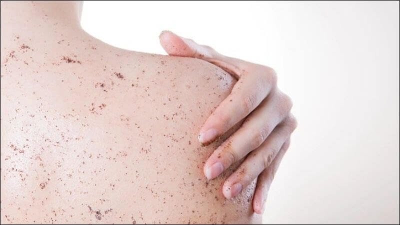 Bờ vai của một người với tay đặt lên, da có các hạt nhỏ, có thể là kem tẩy tế bào chết, nền trắng.