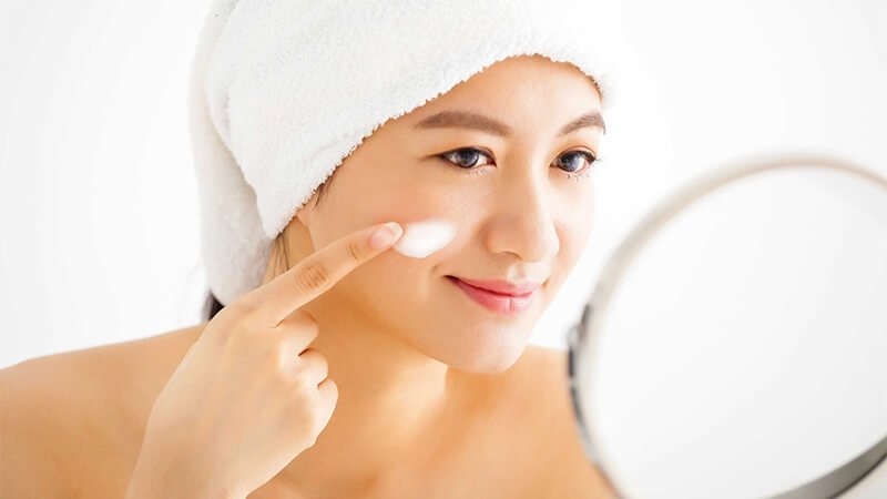 Người phụ nữ quấn khăn tắm, bôi kem dưỡng da lên má, nhìn vào gương với nụ cười nhẹ.
