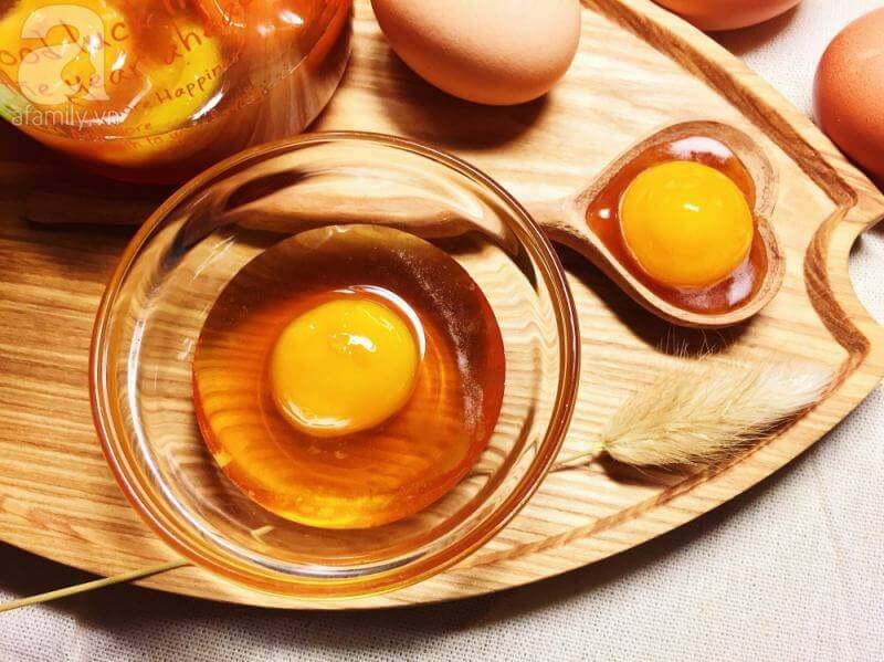 Trứng gà sống trong bát thủy tinh trên khay gỗ, bên cạnh là một chiếc thìa và vài quả trứng nguyên vỏ.