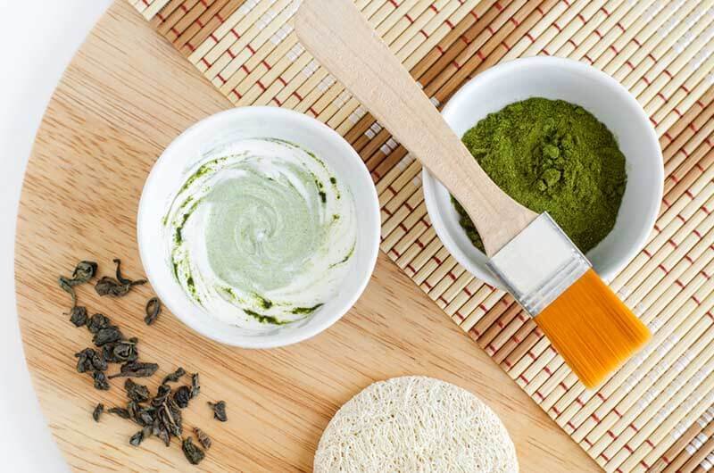 Bột trà xanh và mặt nạ tự chế trong bát, cọ quét, miếng bọt biển và lá trà khô trên nền tre và gỗ.